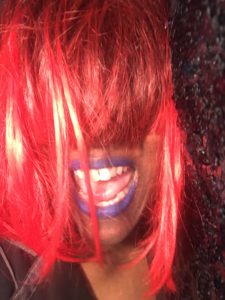 Marya Errin Jones in, "Ask An American," wearing a red wig and purple lipstick