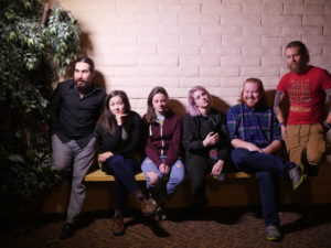 Part of Blackout Theatre Company pose in a line on a bench, situated in front of a blank white wall, with a tree to the left.